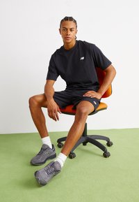 Black cotton t-shirt with a small white logo, black shorts, and gray athletic shoes with mesh detailing, sitting on an orange chair. Green flooring.