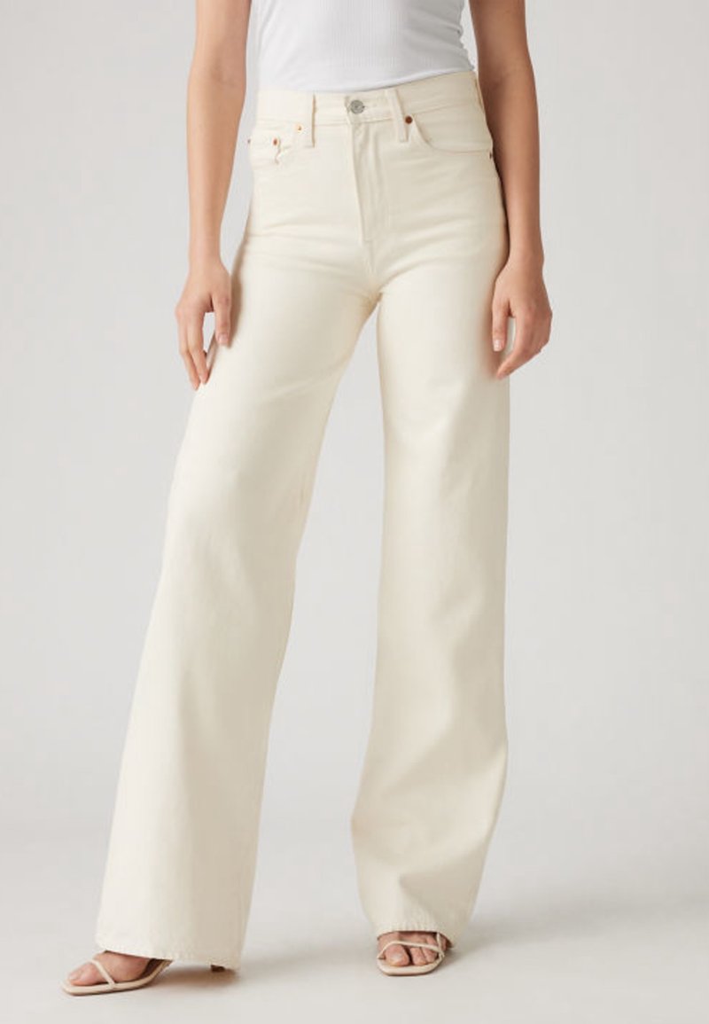 Femme portant un jean taille haute à jambes larges de couleur blanc cassé, associé à un top blanc sans manches et des sandales beiges à brides.