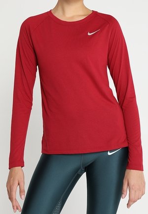 Žena oblečená v červeném dlouhém rukávu Nike a tmavě tyrkysových legínách Nike, stojící s jednou rukou uvolněnou a druhou mírně pokrčenou.