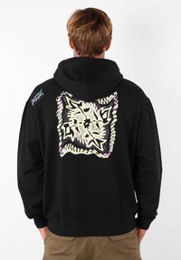 Haut à capuche noir avec un grand motif géométrique pastel au dos. Il dispose d'une poche kangourou et de poignets côtelés. Tissu texturé et lourd.