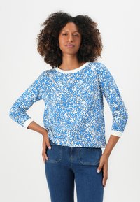 Blå långärmad topp med ett vitt abstrakt mönster och kontrasterande vit krage. Mjuk tyg; avslappnad passform; kombinerad med mörka denimjeans.