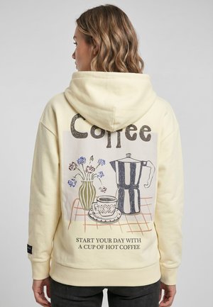 Vrouw met golvend haar draagt een crèmekleurige hoodie met een koffiepot, kopje, bloemen en tekst die hete koffie op de achterkant promoot.