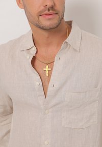 Beige linned button-up skjorte med lomme, let åben krave og en guldkorshalskæde på en ensfarvet baggrund.