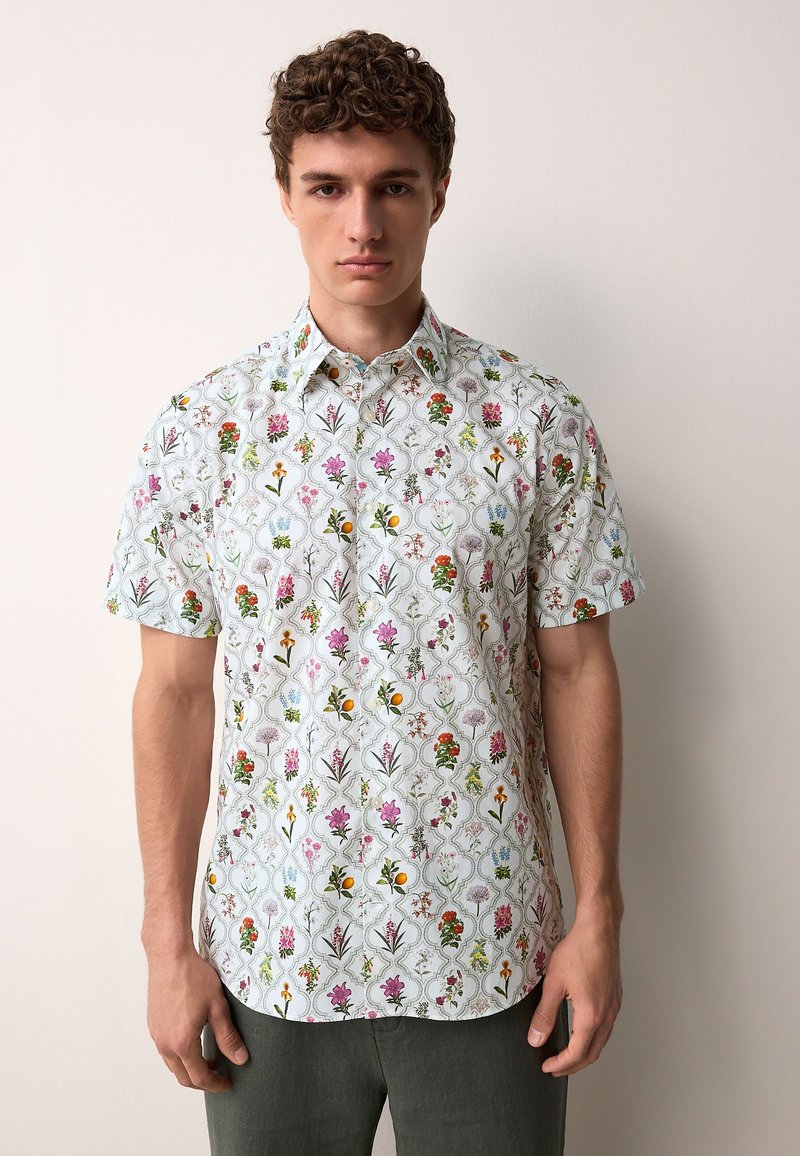 Chemise à manches courtes avec une base blanche, présentant un motif floral de différentes couleurs, dont le rose et le vert, et un col boutonné.