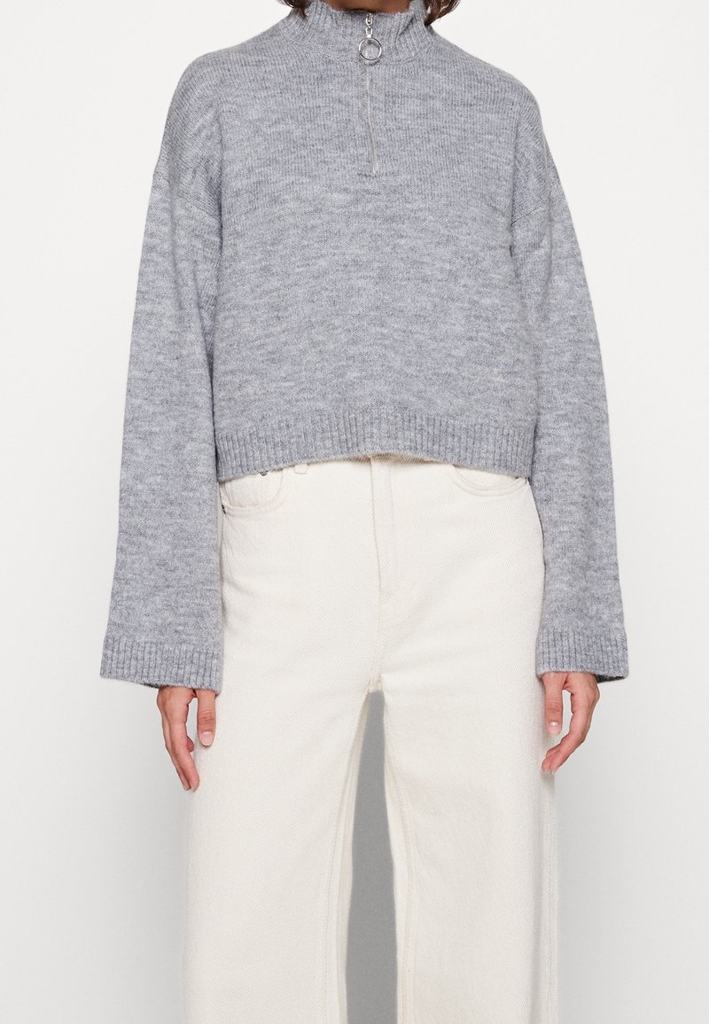 Szary, dziany krótki sweter z wysokim kołnierzem i zamkiem błyskawicznym z przodu. Ściągacze na mankietach i dolnym brzegu. W połączeniu z szerokimi, off-white dżinsami.