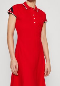 Rode polo jurk met korte mouwen, witte en zwarte gestreepte accenten op de mouwen en kraag, knoopsluiting aan de voorkant, zachte stof, knielang.