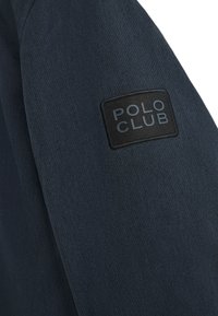 Giacca navy in tessuto texturizzato; presenta una toppa di stoffa nera con la scritta "POLO CLUB" sulla manica.