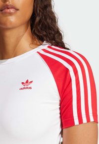 Haut de sport rouge et blanc, en tissu extensible. Présente trois rayures rouges sur les manches, avec un petit logo Adidas rouge sur la poitrine.