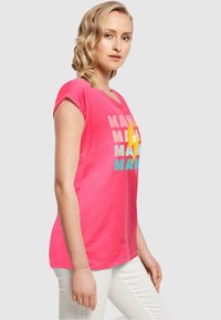 Lyserød bomulds T-shirt med korte ærmer, der har farverig tekst. Designet har et legesygt grafisk element i gul. Bæret med hvide bukser.
