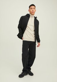 Chaqueta acolchada negra con capucha, suéter beige con cremallera y jeans negros holgados. El calzado incluye zapatillas oscuras con suela texturizada.