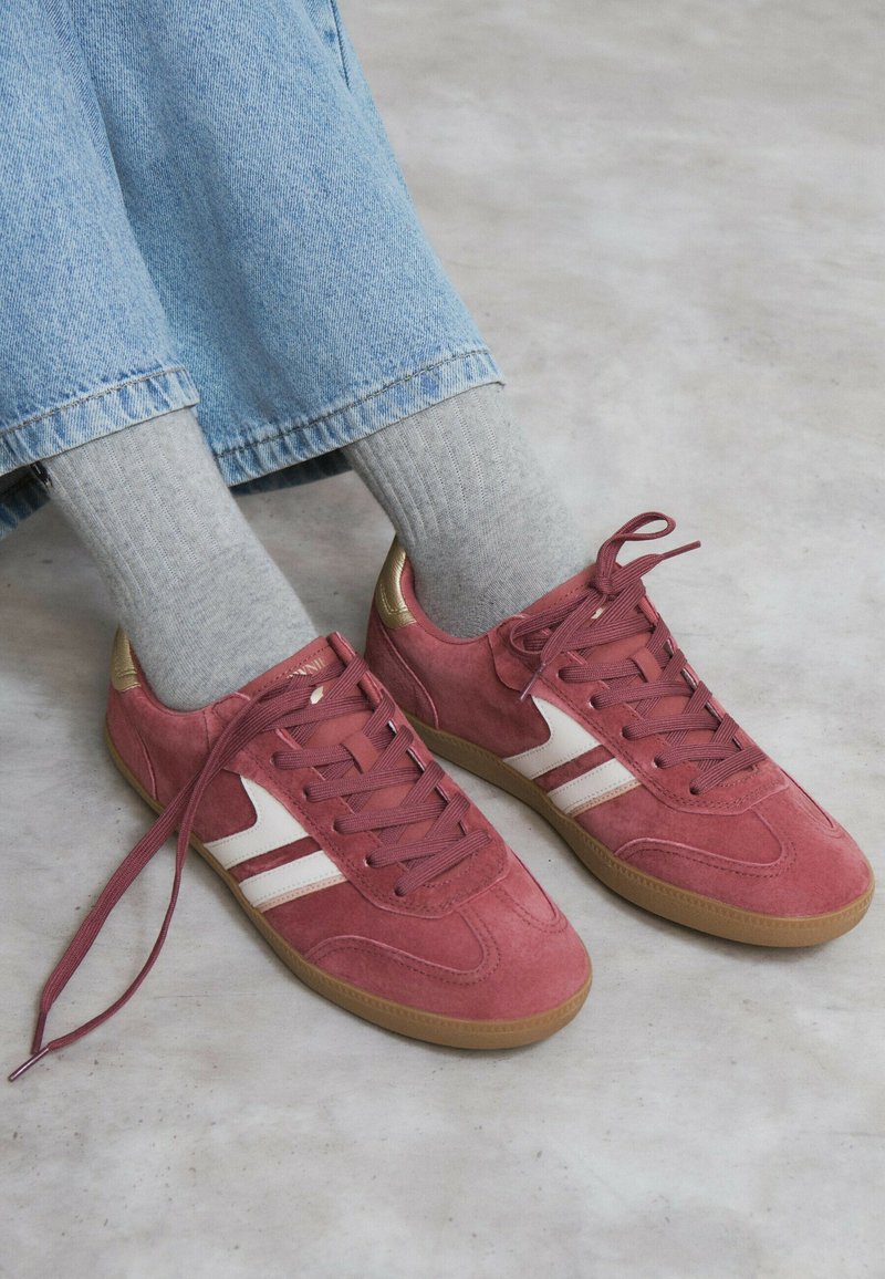 Persona con vaqueros azul claro, calcetines grises y zapatillas de ante rosa con rayas blancas y suelas de goma, de pie sobre un suelo claro.