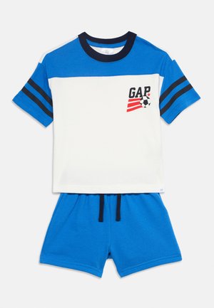 Chemise bleu et blanc à manches courtes avec col noir et rayures, logo de football "GAP" rouge et noir, associée à un short bleu à cordon.
