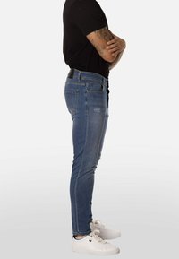 Vaqueros azul denim de corte ajustado, con un diseño clásico de cinco bolsillos, sutil desgastado y una etiqueta negra en la cinturilla trasera.