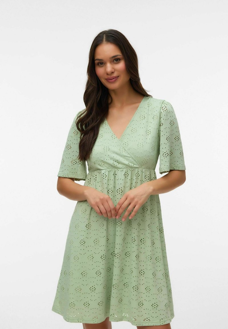 Pruunide pikkade juustega naine, kes kannab helesinist lühikeste varrukatega kleiti silmaaugumustriga, seisab tavalise valge tausta ees.