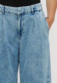 Lyseblå denimbukser med avslappet passform, med folder, beltehemper og sidelommer. Stoffet har et litt vaskeskyet utseende.