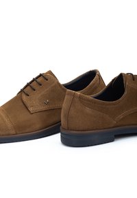 Zapatos de ante marrón con cordones, con punta redondeada, textura suave, detalles de costura sutiles, y con un interior negro acolchado y suela de goma.