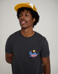 Fekete pamut rövid ujjú póló színes mellkasi grafikával. Egy sárga hálós hátú sapkát enyhén ferdén viselnek.