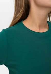 Teal rebrasti majica s okruglim izrezom. Tkanina izgleda mekano s glatkom teksturom i prilagođenim dizajnom koji naglašava slim oblik.