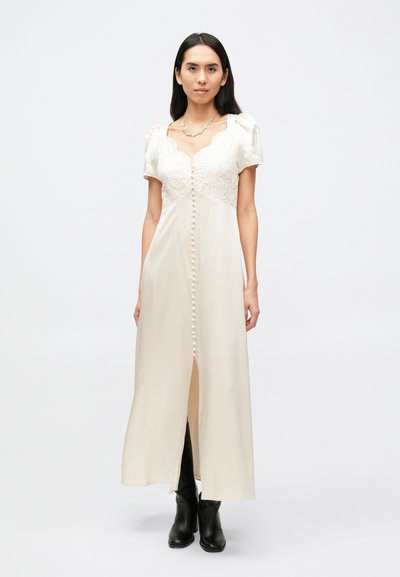 Robe en satin crème avec des détails en dentelle, des manches courtes bouffantes et des boutons à l'avant. Présente une fente au centre. Style complété par des bottes noires.