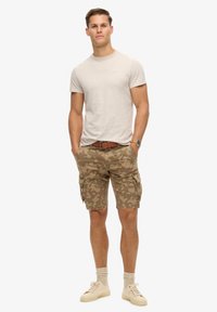 Beige katoenen t-shirt gecombineerd met camouflage cargoshorts in bruin en groen, met zijzakken. Neutrale schoenen maken de look compleet.