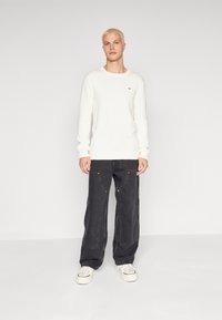 Suéter blanco de punto acanalado con cuello redondo, combinado con pantalones cargo negros holgados que tienen cremalleras. El modelo lleva zapatillas blancas.