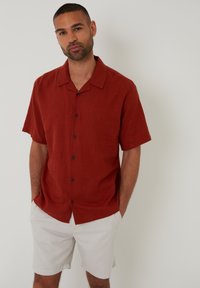 Rød kortærmet skjorte i let materiale med krave og sorte knapper foran. Kombineret med lyse shorts.
