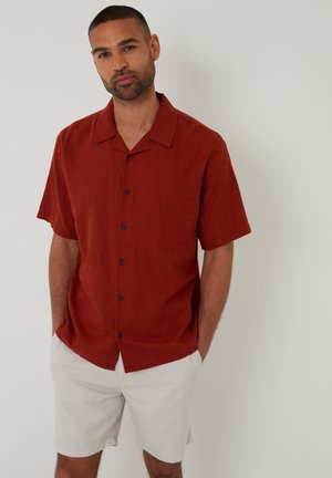 Camisa roja de manga corta en tejido ligero con cuello, que presenta botones negros en el frontal. Combinada con pantalones cortos claros.