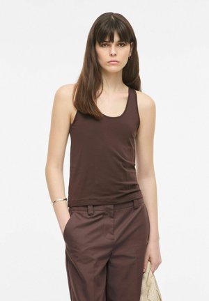 Femme aux cheveux bruns raides, portant un haut marron sans manches et un pantalon assorti, debout avec une main dans la poche et tenant un tissu léger.
