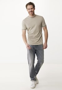 T-shirt beige chiaro con rifiniture grigie a contrasto sul collo e sulle maniche, abbinata a jeans grigi e sneakers bianche.