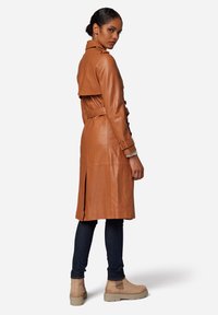 Tan leather trench coat with a double-breasted design, belted waist, and shoulder flaps. Mid-length with a center back vent. Casual boots visible.