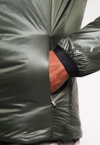 Oliven grøn jakke lavet af letvægts, skinnende materiale. Har elastiske manchetter, en lynlås-lomme og en tætsiddende design.