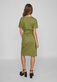 Olivově zelené tričkové šaty, délka ke kolenům, měkká látka, krátké rukávy, překroucený detail na zádech, v kombinaci s černými platformovými sandály.