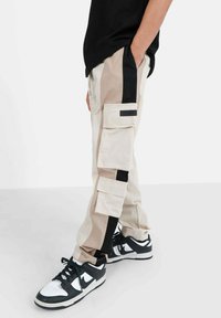 Beige cargobroeken met zwarte zijkanten, meerdere zakken en een contrasterend textuurpaneel. Gecombineerd met zwart-witte sneakers.