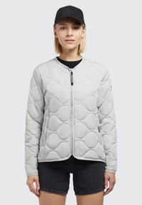 Lichtgrijze gewatteerde jas met een ronde hals, vooraan met rits en zijzakken. G gedragen over een zwarte shirt en gecombineerd met zwarte shorts.