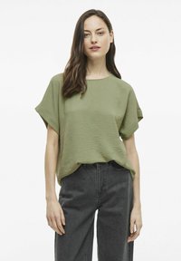 Alyvuogių žalios spalvos trumpas rankovėmis marškinėliai su tekstūruotu paviršiumi, spalva derinama su juodais aukštu juosmeniu džinsais. Paprastas, laisvas siluetas su apvalia iškirpte.