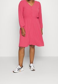 Ljusrosa klänning med V-hals, knapptyg och långa ärmar. Har en rynkad midja och en svävande kjol. Bärs med vita sneakers.