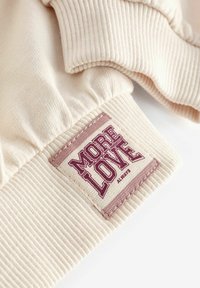 Cremefarbener Baumwollmischgewebe mit gerippter Textur; ausgestattet mit einem pinkfarbenen aufgestickten Etikett mit der Aufschrift "MEHR LIEBE IMMER."