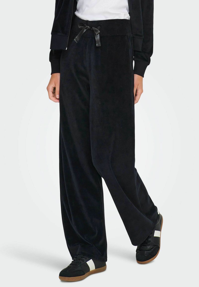 Pantalon large en velours noir avec taille élastique et cordon de serrage en satin noir, assorti à des baskets noires avec des accents blancs.