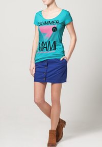 Camiseta de color teal con un gráfico que dice "VERANO EN MIAMI" y mangas cortas, combinada con una falda azul de botón frontal y botas de tobillo marrones.