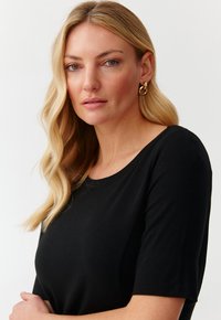 Mustat lyhythihaiset paidat, joissa on pyöreä kaula-aukko. Sileä rakenne ja vartalonmyötäinen muotoilu. Yhdistetty suurten kultaketjujen korvakorujen kanssa. Neutraali tausta.