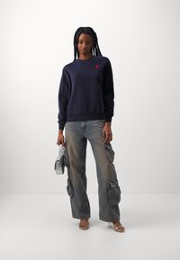 Felpa navy con scollo tondo e logo a forma di cuore rosso; abbinata a jeans cargo sbiaditi con grandi tasche laterali; borsa argentata e sandali trasparenti.