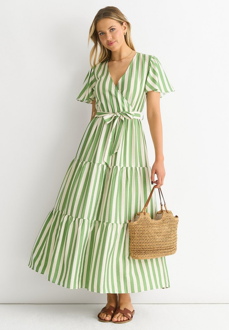Green and white striped maxi dress with a wrap design, short flutter sleeves, a tie belt, and tiered skirt; accessorized with a woven bag.