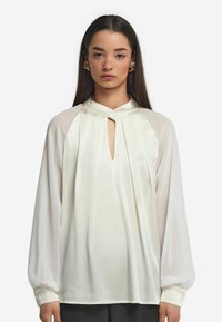 Weiße Bluse mit hohem Kragen, Falten an der Vorderseite und langen, gepufften Ärmeln. Glatte Textur mit Schlüsselspalten-Ausschnitt.