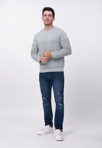 Pull-over gris clair avec un motif texturé, associé à un jean bleu usé et des baskets blanches. Col rond simple, manches longues.
