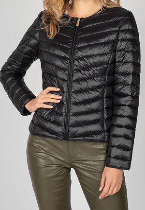 Femme portant une veste noire matelassée zippée à manches longues et un pantalon en cuir vert olive, main posée sur la taille.