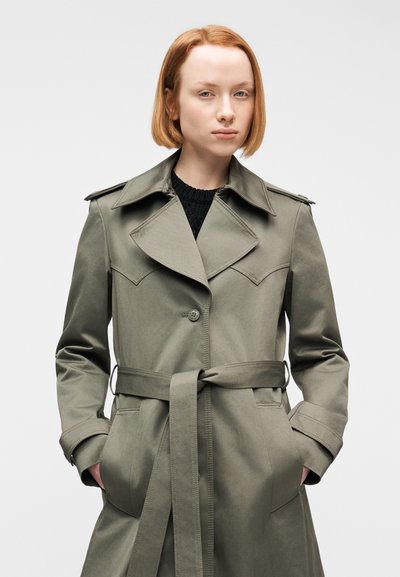 Νέα γυναίκα με κοντά κόκκινα μαλλιά, που φοράει γκρι παλτό με ζώνη πάνω από μαύρο πουλόβερ, με τα χέρια στις τσέπες του παλτού, πάνω σε απλό λευκό φόντο.