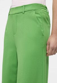 Pantalones verdes a medida con frente plano, bolsillos laterales y una tela texturada. La cinturilla es ajustable para mayor comodidad.