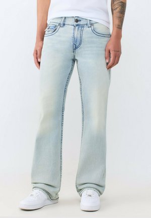 Persoon die lichtblauwe wijde jeans draagt, witte sneakers, een wit overhemd, met een tatoeage zichtbaar op de rechter onderarm.