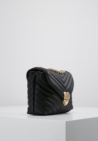 Svart vadderad läderhandväska med chevronmönster, guldhjätratformad spänne och guldkedjeaxelrem, placerad på en vit pelare.