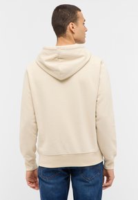 Beżowa bluza z kapturem o miękkiej fakturze, luźnym kroju, ściągaczach na rękawach i kieszeni typu kangurka, zestawiona z niebieskimi dżinsami.
